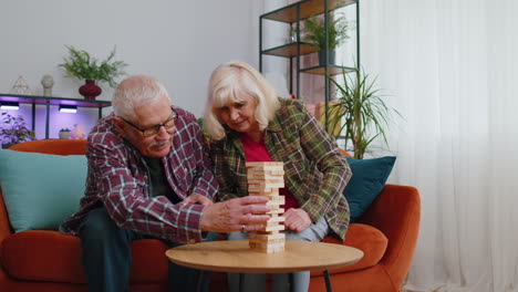 Elderly-old-grandparents-man-woman-playing-in-blocks-wooden-tower-build-board-game-on-table-at-home