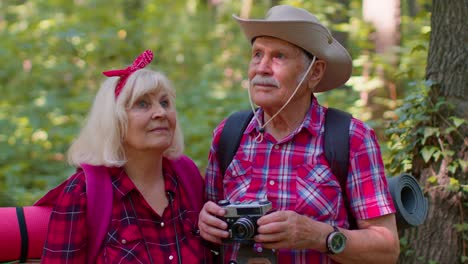 Senior-old-grandmother-grandfather-tourists-walking-with-backpacks-taking-photos-with-camera-in-wood