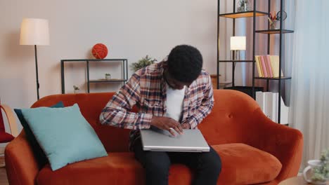 Afroamerikanischer-Mann-Sitzt-Auf-Dem-Sofa-Und-öffnet-Seinen-Laptop-PC,-Während-Er-Im-Wohnzimmer-Zu-Hause-Online-Mit-Der-Arbeit-Beginnt