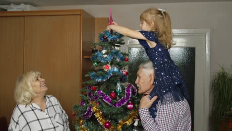 Una-Niña-Pequeña-Y-Linda-Con-Una-Familia-De-Abuelos-Mayores-Decorando-Un-árbol-De-Navidad-Artificial-En-Casa
