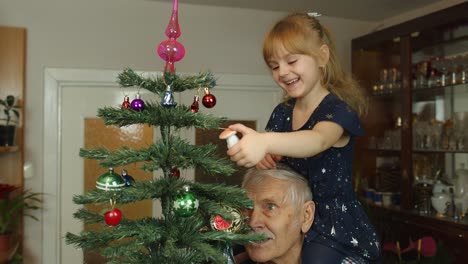 Girl-kid-with-senior-grandpa-decorating-artificial-Christmas-tree-with-ornaments-and-toys-at-home