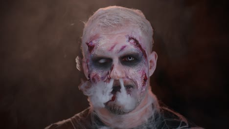 Sinister-man-Halloween-zombie-with-bloody-face-blows-smoke-from-nose-and-mouth,-making-air-fly-kiss