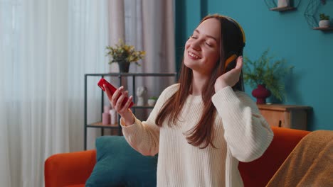 Pretty-Caucasian-young-woman-in-headphones-listening-music-dancing,-singing-in-living-room-at-home