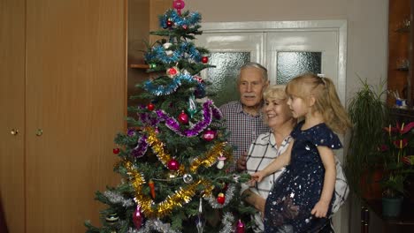 Senior-grandfather,-grandmother-and-granddaughter-child-lights-garlands-on-artificial-Christmas-tree