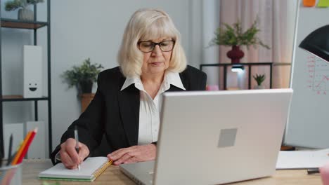 Senior-mature-older-business-office-woman-elearning-online-writing-notes-from-laptop,-live-webinar