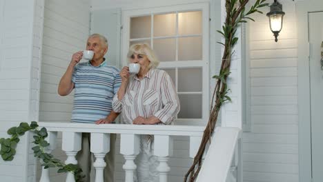 Senior-elderly-Caucasian-couple-drinking-coffee,-embracing-in-porch-at-home.-Mature-happy-family