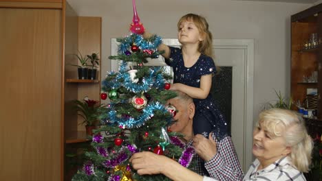 Children-girl-with-elderly-couple-grandparents-decorating-artificial-Christmas-pine-tree-at-home
