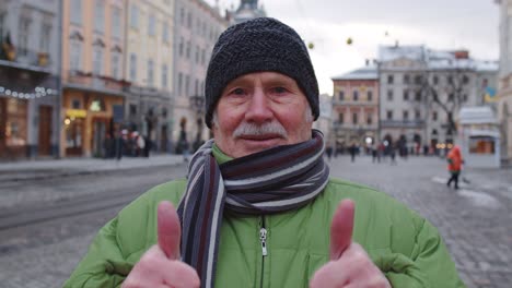 Stylish-old-senior-man-tourist-smiling,-showing-thumbs-up-in-winter-city-center-of-Lviv,-Ukraine