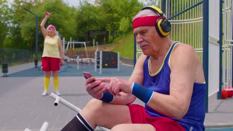 Senior-old-sportsman-grandfather-with-headphones-listening-music-from-mobile-phone-on-playground