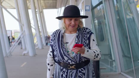 Senior-tourist-grandmother-woman-walking-on-international-airport-hall,-using-mobile-phone,-texting