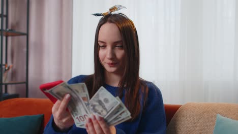 Smiling-happy-young-woman-holding-cash-money-and-mobile-phone-calculate-domestic-bills-at-home