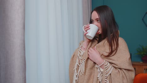 Young-adult-brunette-girl-in-warm-white-sweater-drink-hot-beverage-mug-relaxing-at-home-near-window