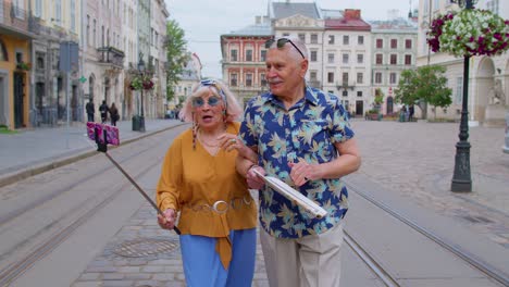Senior-old-tourists-man-with-woman-walking-in-city-with-smartphone-on-selfie-stick-having-video-call