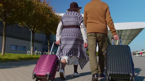 Senior-husband-wife-retirees-tourists-go-to-airport-terminal-for-boarding-with-luggage-on-wheels