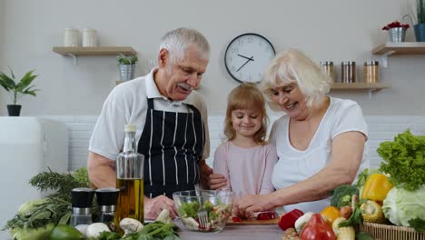 Senior-couple-grandmother-and-grandfather-in-kitchen-feeding-granddaughter-child-with-chopped-pepper
