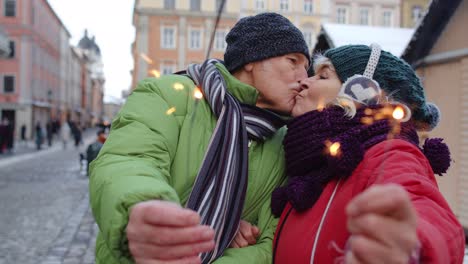Senior-couple-holding-sparklers-bengal-lights-enjoying-Christmas-eve,-making-kiss-in-winter-city