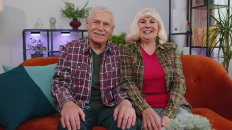 Happy-old-senior-elderly-family-couple-hugging,-laughing,-smiling-looking-at-camera-at-home-sofa