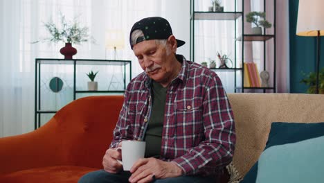 Älterer,-Stilvoller-Großvater,-Der-Auf-Der-Couch-Sitzt-Und-Zu-Hause-Entspannt-Und-Tee-Oder-Kaffee-Trinkt