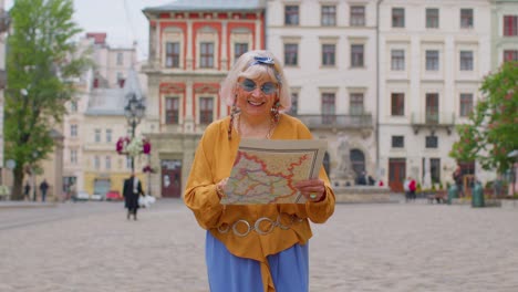 Elderly-stylish-tourist-granny-woman-walking-along-street,-looking-for-way-using-paper-map-in-city