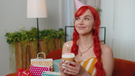 Happy-redhead-girl-celebrating-birthday-party,-makes-wish-blowing-burning-candle-on-small-cupcake
