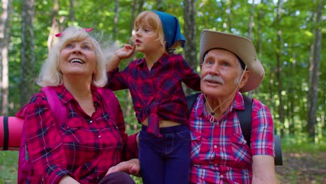 Old-senior-grandparents-couple-tourists-hikers-resting-in-forest-with-granddaughter-in-summer-wood