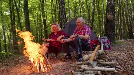 Happy-senior-man-and-woman-talking,-cooking-frying-sausages-over-campfire-in-evening-wood,-camping