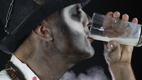 Scary-guy-in-carnival-costume-of-Halloween-skeleton-looking-at-camera,-drinks-milk-from-a-glass