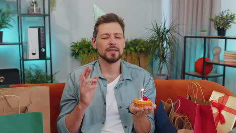 Happy-young-adult-man-celebrating-birthday-party,-makes-wish-blowing-burning-candle-on-small-cupcake