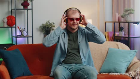 Overjoyed-young-man-in-wireless-headphones-dancing,-singing-on-cozy-couch-in-living-room-at-home