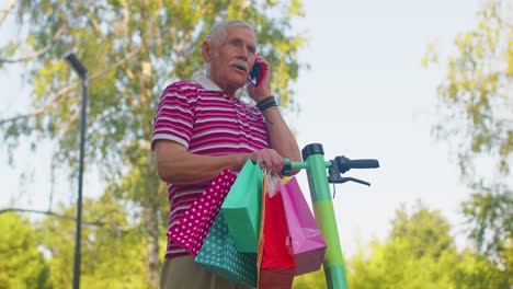 Anciano-Abuelo-Hombre-En-Scooter-Eléctrico-Con-Coloridas-Bolsas-De-Compras-Hablando-Por-Teléfono-Móvil