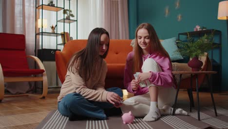Girls-friends-siblings-sitting-on-floor-and-take-turns-dropping-dollar-banknote-into-piggy-bank
