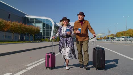 Stylish-retired-family-couple-granny-grandfather-walking-with-luggage-suitcase-bags-to-airport-hall