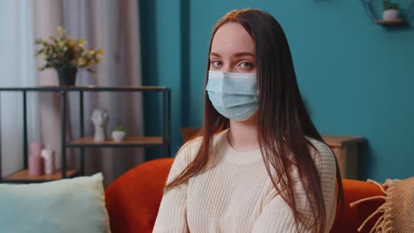Portrait-of-young-sick-girl-wearing-medical-protection-mask-sitting-in-living-room-looking-at-camera