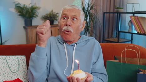 Happy-elderly-old-man-celebrating-birthday-party,-makes-wish-blowing-burning-candle-on-small-cupcake