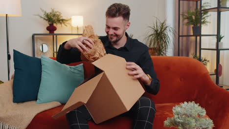 Happy-young-guy-shopper-unpacking-cardboard-box-delivery-parcel-online-shopping-purchase-at-home