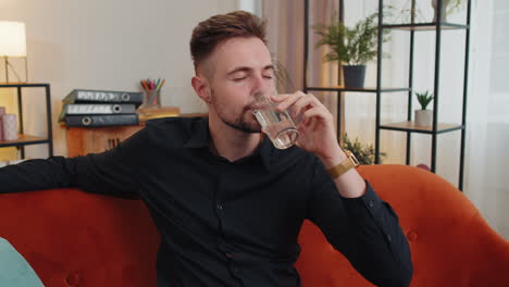 Ein-Durstiger-Junger-Kerl-Sitzt-Zu-Hause-Und-Hält-Ein-Glas-Natürliches-Aqua,-Trinkt-Stilles-Wasser-Und-Trinkt-Schlucke