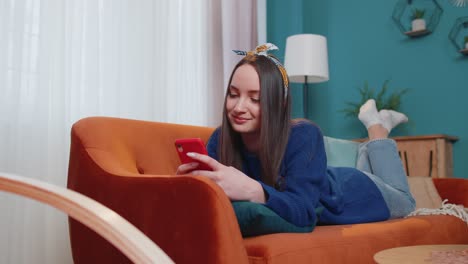 Joyful-young-adult-woman-lying-on-sofa-using-smartphone-watching-video,-online-shopping,-tapping