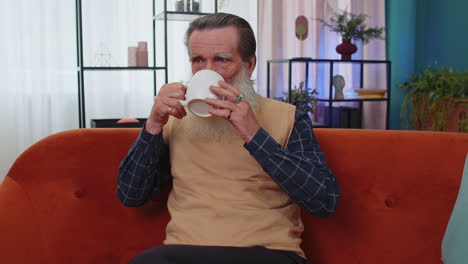Attractive-smiling-senior-old-grandfather-man-drinking-a-cup-of-coffee-or-herbal-tea-at-home-couch