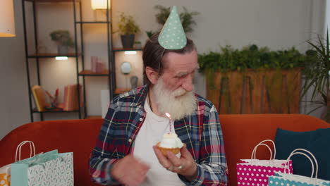 Happy-elderly-old-man-celebrating-birthday-party,-makes-wish-blowing-burning-candle-on-small-cupcake