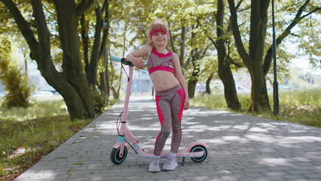 Athletic-fit-young-girl-rides-on-electric-scooter,-standing-near-bike-looking-at-camera-in-park