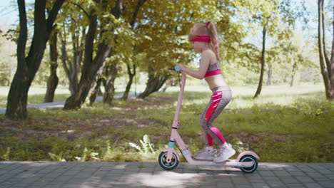 Young-athletic-fit-blonde-girl-rides-traveling-on-electric-scooter-on-road-in-park-on-sunny-day