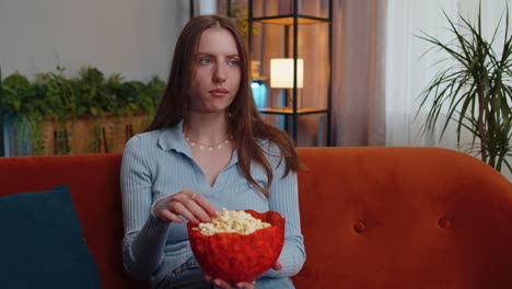 Woman-sitting-on-couch-eating-popcorn-and-watching-interesting-TV-serial,-sport-game-online-at-home