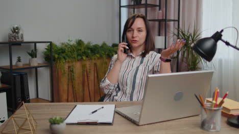 Young-woman-freelancer-answering-to-client-on-mobile-phone-call,-using-laptop-computer,-home-office