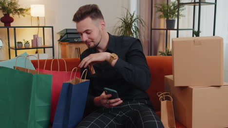 Happy-shopaholic-young-guy-sitting-with-shopping-bags-making-online-payment-with-credit-card