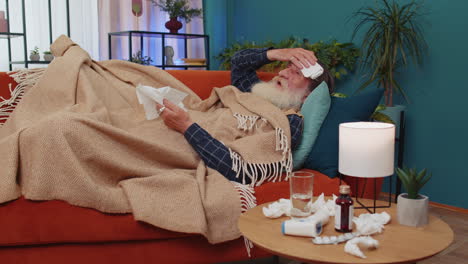 Sick-ill-senior-man-suffering-from-cold-or-allergy-lying-on-home-sofa-sneezes-wipes-snot-into-napkin