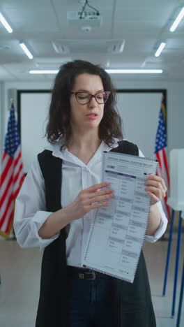 American-woman-speaks-on-camera,-shows-paper-bulletin,-calls-for-voting.-National-Elections-Day-in-the-United-States.-Voting-booths-at-polling-station.-Concept-of-civic-duty-and-patriotism.-Portrait.
