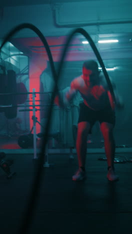 Athletic-man-trains-with-battle-ropes-in-dark-boxing-gym-with-LED-lighting.-Professional-male-boxer-does-cardio-or-endurance-exercises-before-fighting-competition.-Vertical-shot