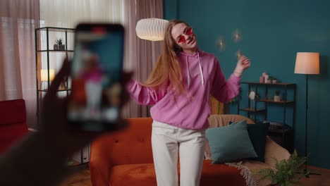 Joyful-blogger-teen-girl-dancing-at-camera-filming-video-using-mobile-phone-at-home-in-living-room