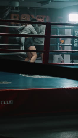 Female-boxer-in-boxing-gloves-practices-fighting-technique-with-male-coach-and-prepares-to-tournament-in-dark-gym.-Athletic-fighter-hits-punching-mitts-on-boxing-ring.-Vertical-shot