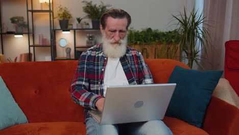 Senior-grandfather-man-sitting-on-sofa-closing-laptop-pc-after-finishing-work-in-living-room-at-home